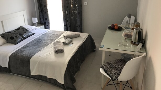 Una cama bien hecha con colcha blanca y gris, un almohadón gris y dos toallas enrolladas. Una mesita con superficie de vidrio sostiene una tetera, un vaso y un plato con frutas. Una silla blanca está colocada junto a la mesa.