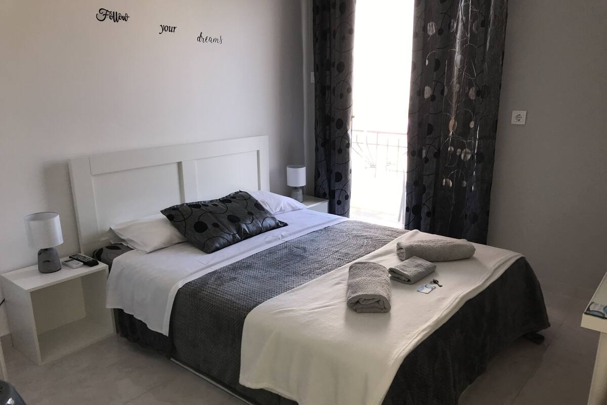 Una cama bien hecha con edredón blanco y gris, una almohada negra y una manta gris. Hay dos mesitas de noche, una con una lámpara y un control remoto, y la otra con una lámpara. La habitación tiene una pared blanca con una cita motivacional y una ventana con cortinas.