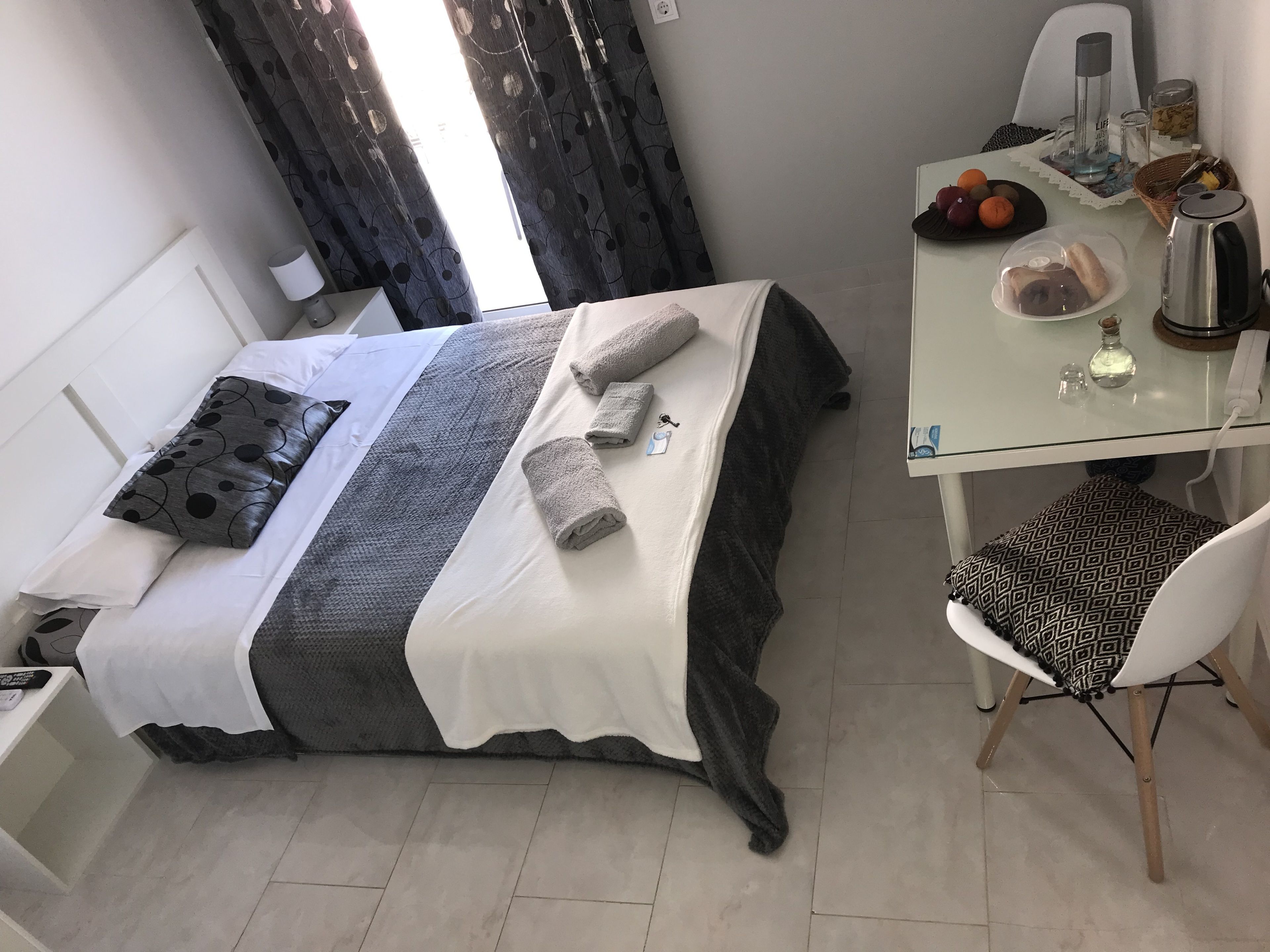 Un lit bien fait, recouvert d’une courtepointe blanche et grise, avec deux oreillers et des serviettes enroulées. Une petite table, sur laquelle se trouvent une bouilloire, un verre et un panier de fruits, est placée à côté du lit.