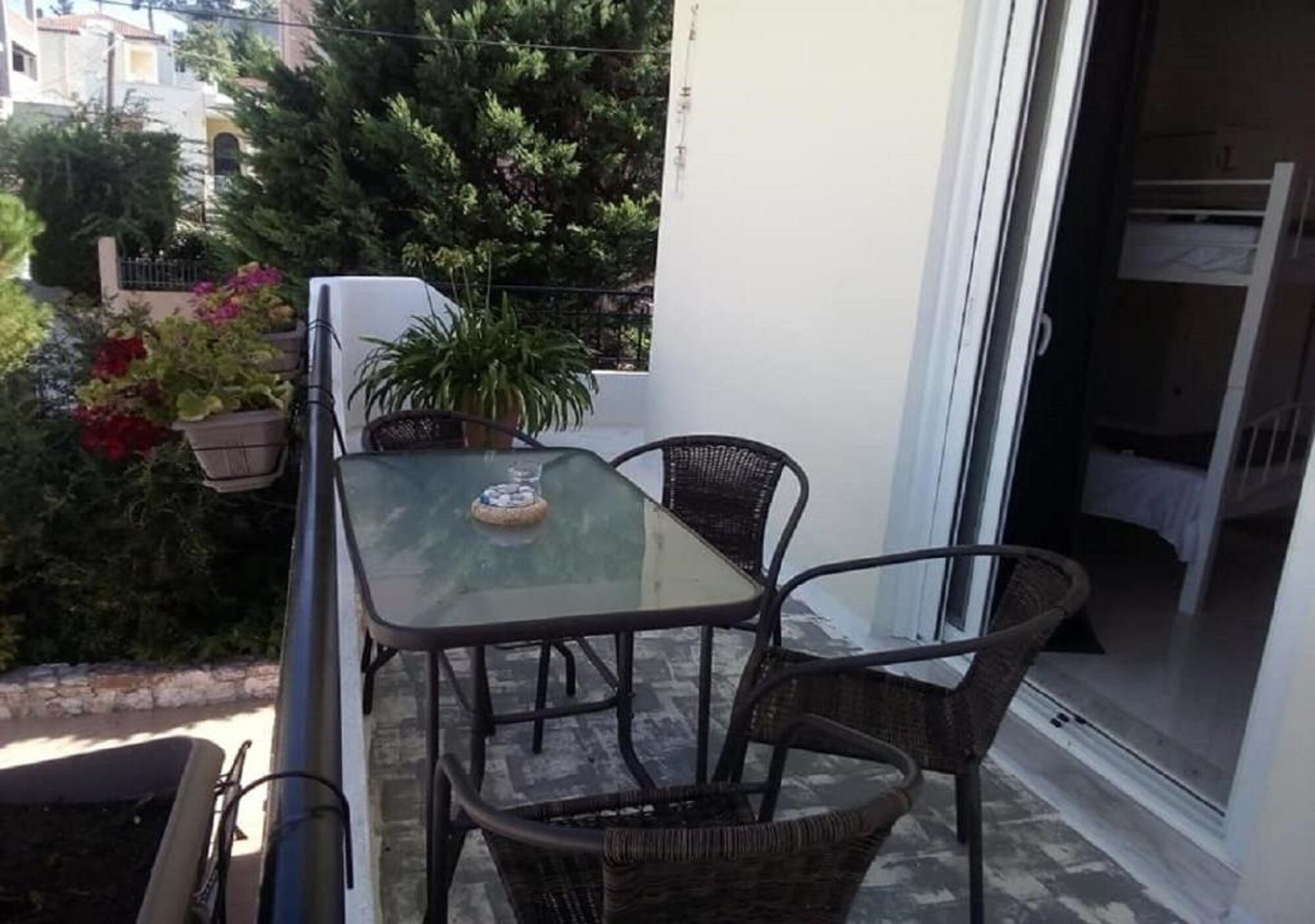 Un balcon avec une table en verre et des chaises en osier, entouré de plantes en pot et offrant une vue sur les bâtiments voisins.