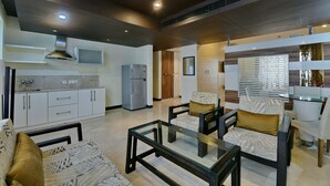 A modern living room with a glass coffee table, patterned sofas, and a kitchen area with white cabinets and a built-in oven. - Hotel Imperial Villa (New Delhi)