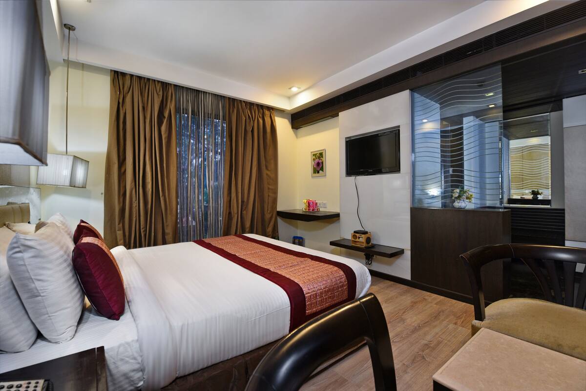 Habitación de hotel con cama, escritorio, silla, televisor y ventana con cortinas.