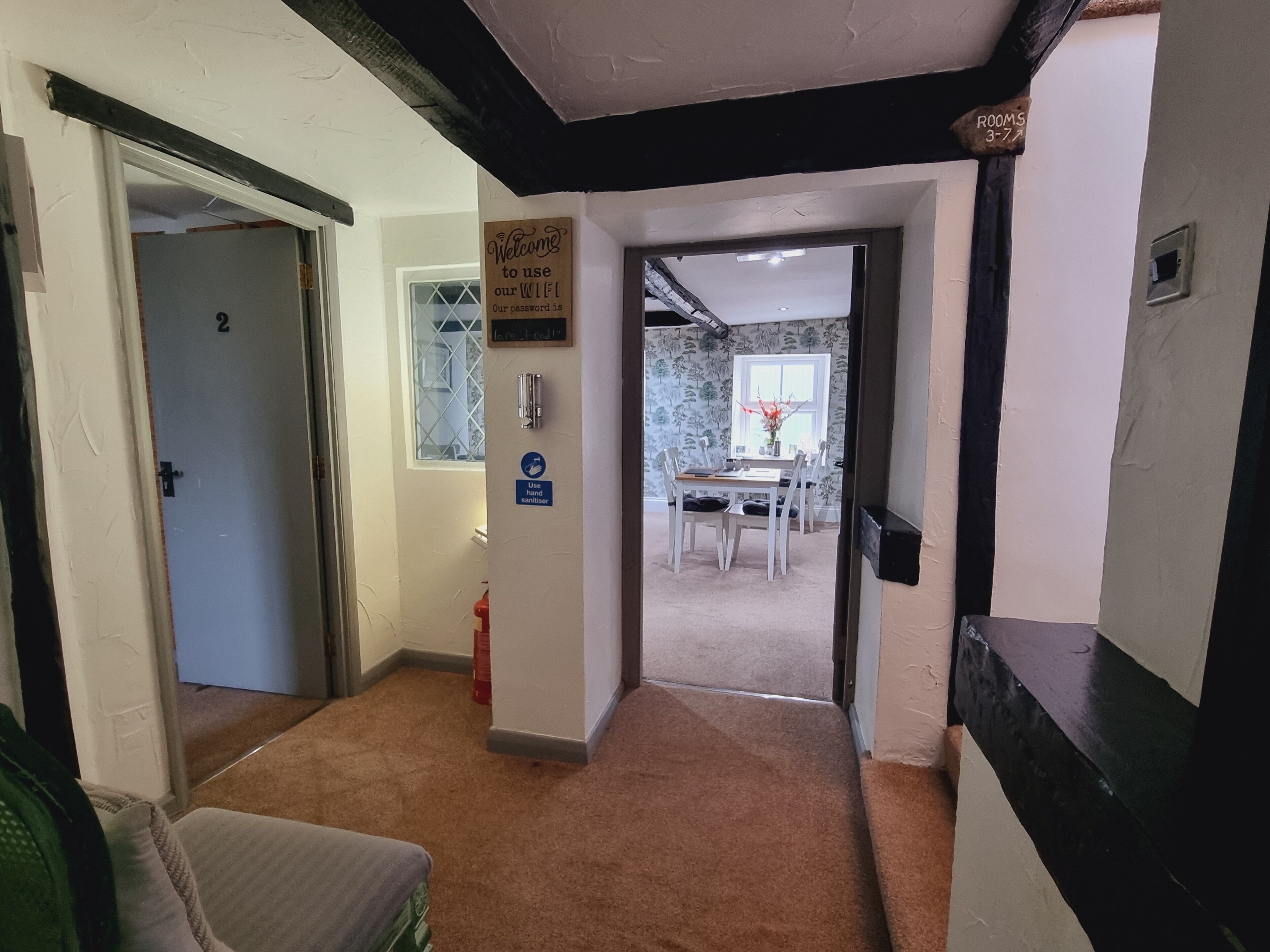 A hallway with a carpeted floor, a sign indicating the use of hand sanitizer, and a view into a dining area with a table and chairs.