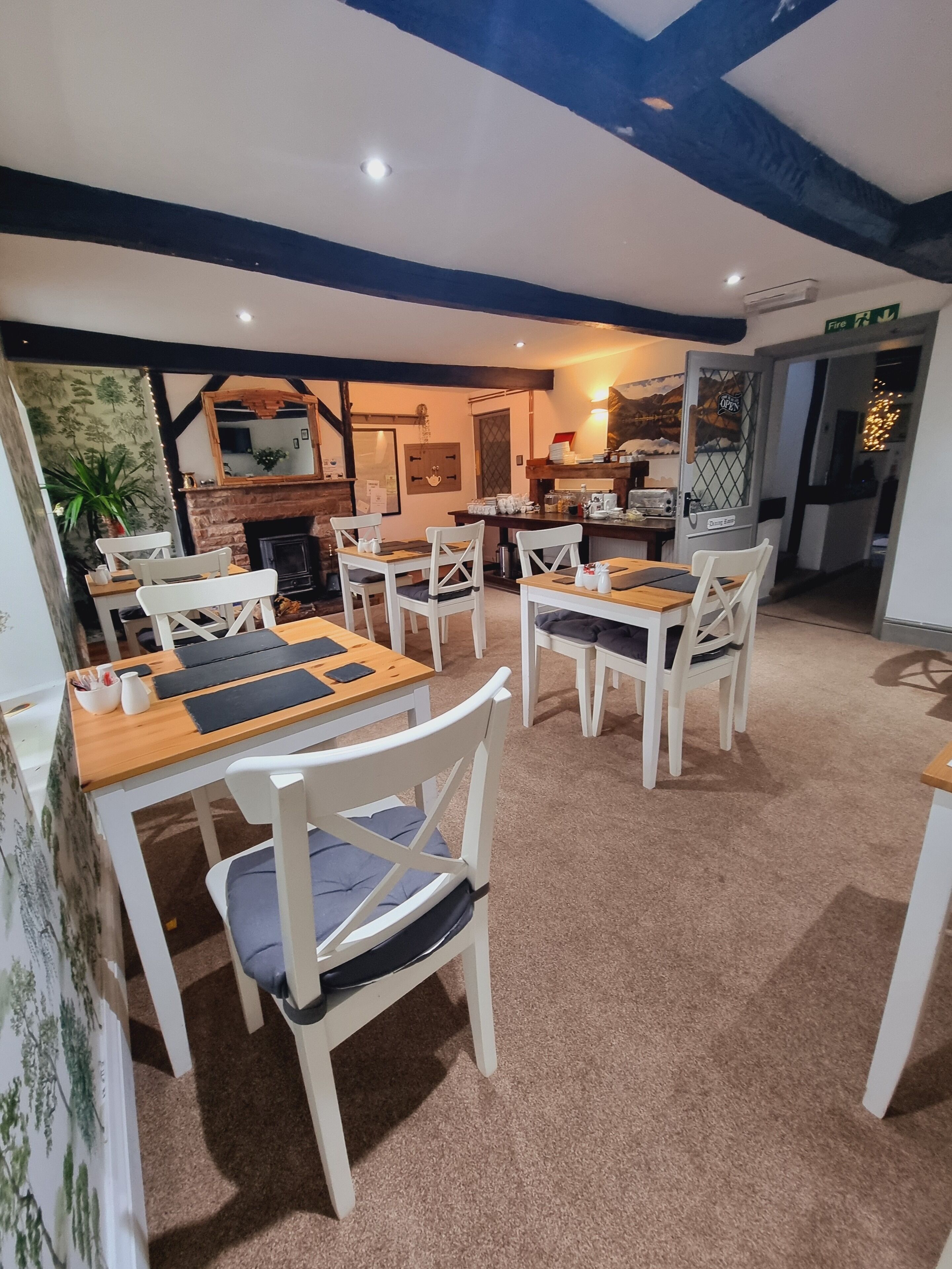 A dining area with white chairs and wooden tables, a fireplace, and a kitchen in the background.