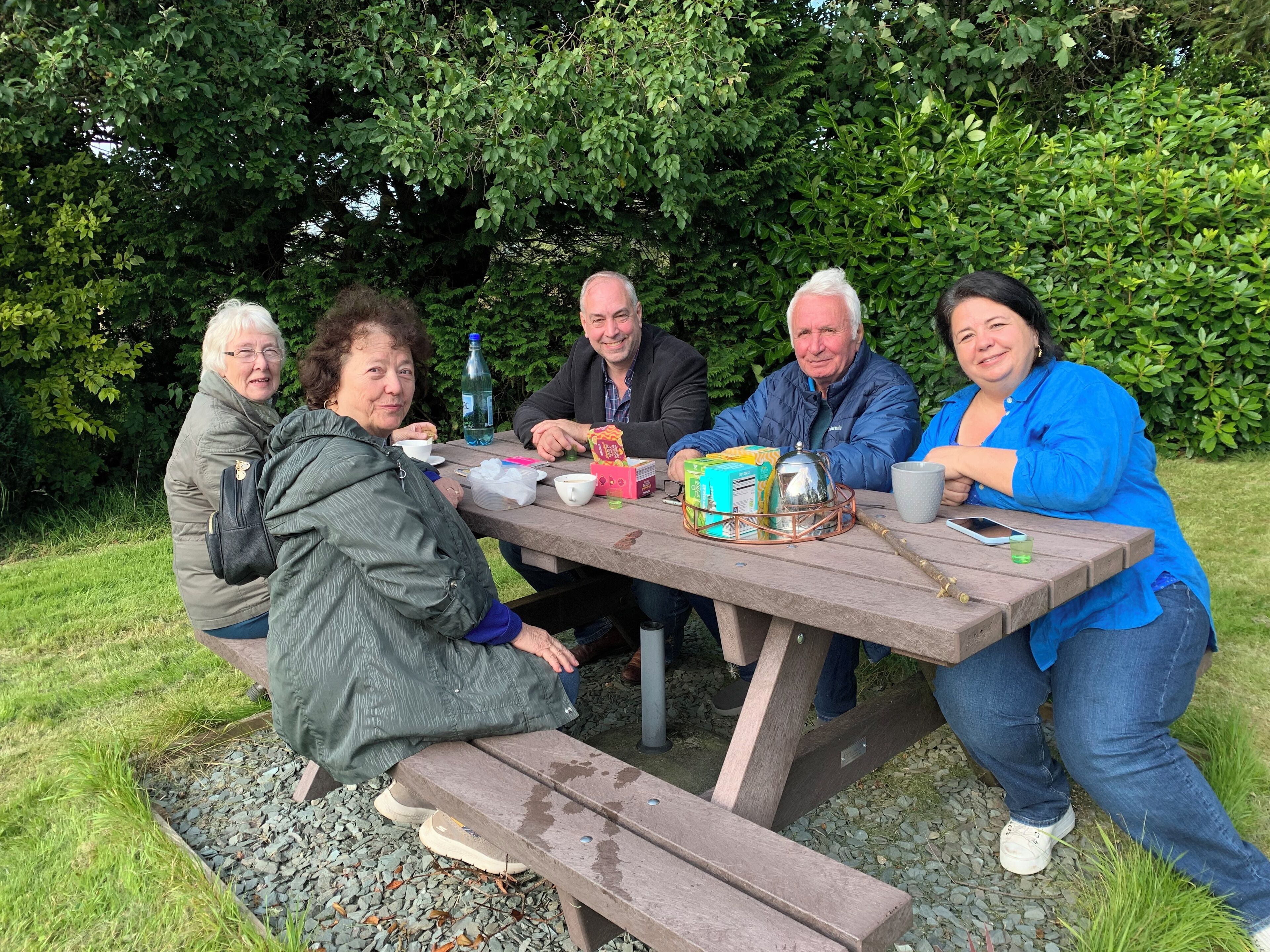 A group of people sitting at an outdoor wooden table with picnic supplies.