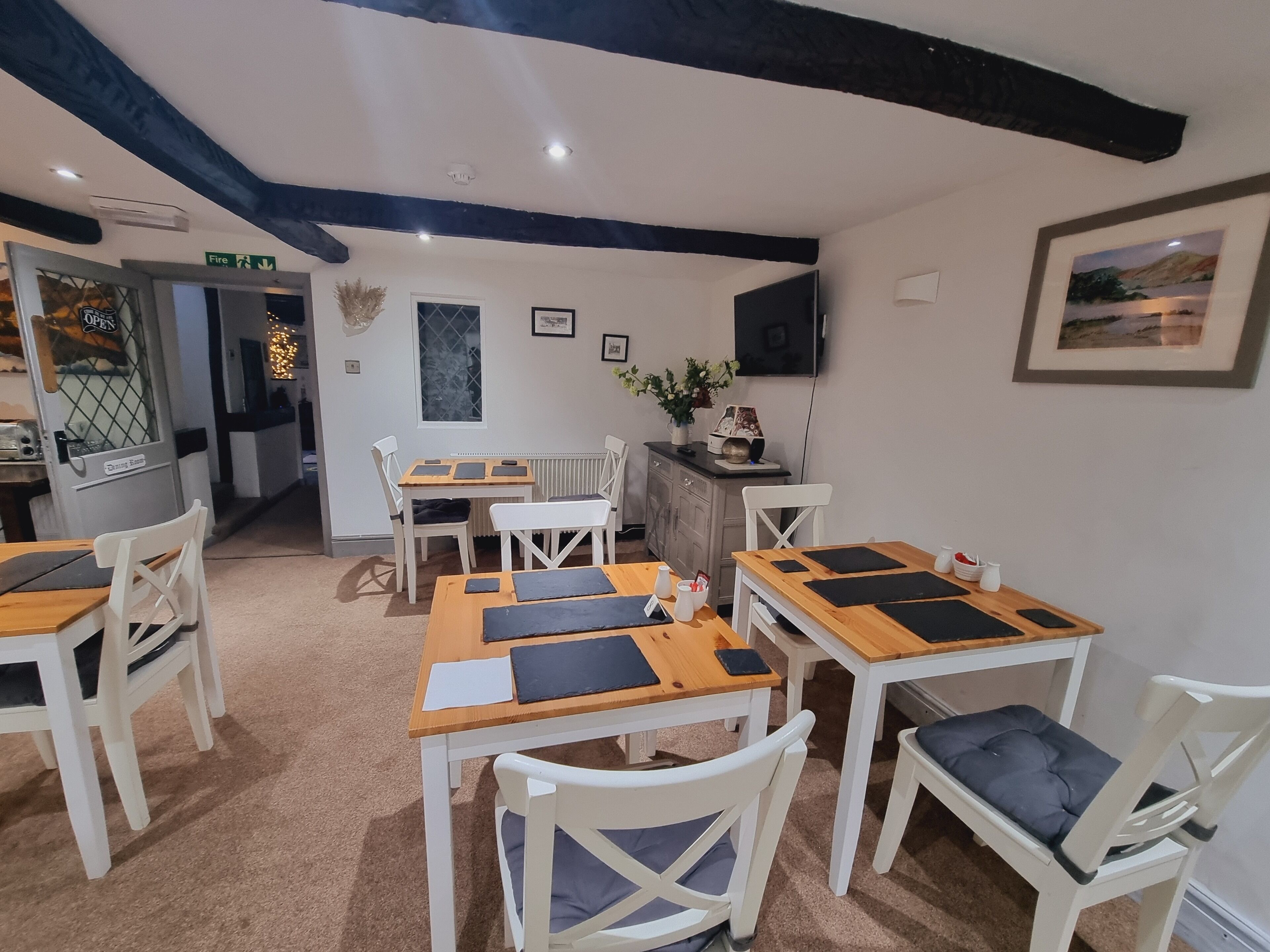 Un espace repas avec des tables en bois et des chaises blanches, un téléviseur fixé au mur et un tableau encadré au-dessus du comptoir.