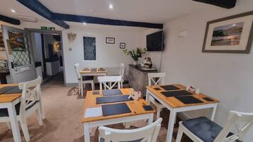 Un espace repas avec des tables en bois et des chaises blanches, un téléviseur fixé au mur et un tableau encadré au-dessus du comptoir.