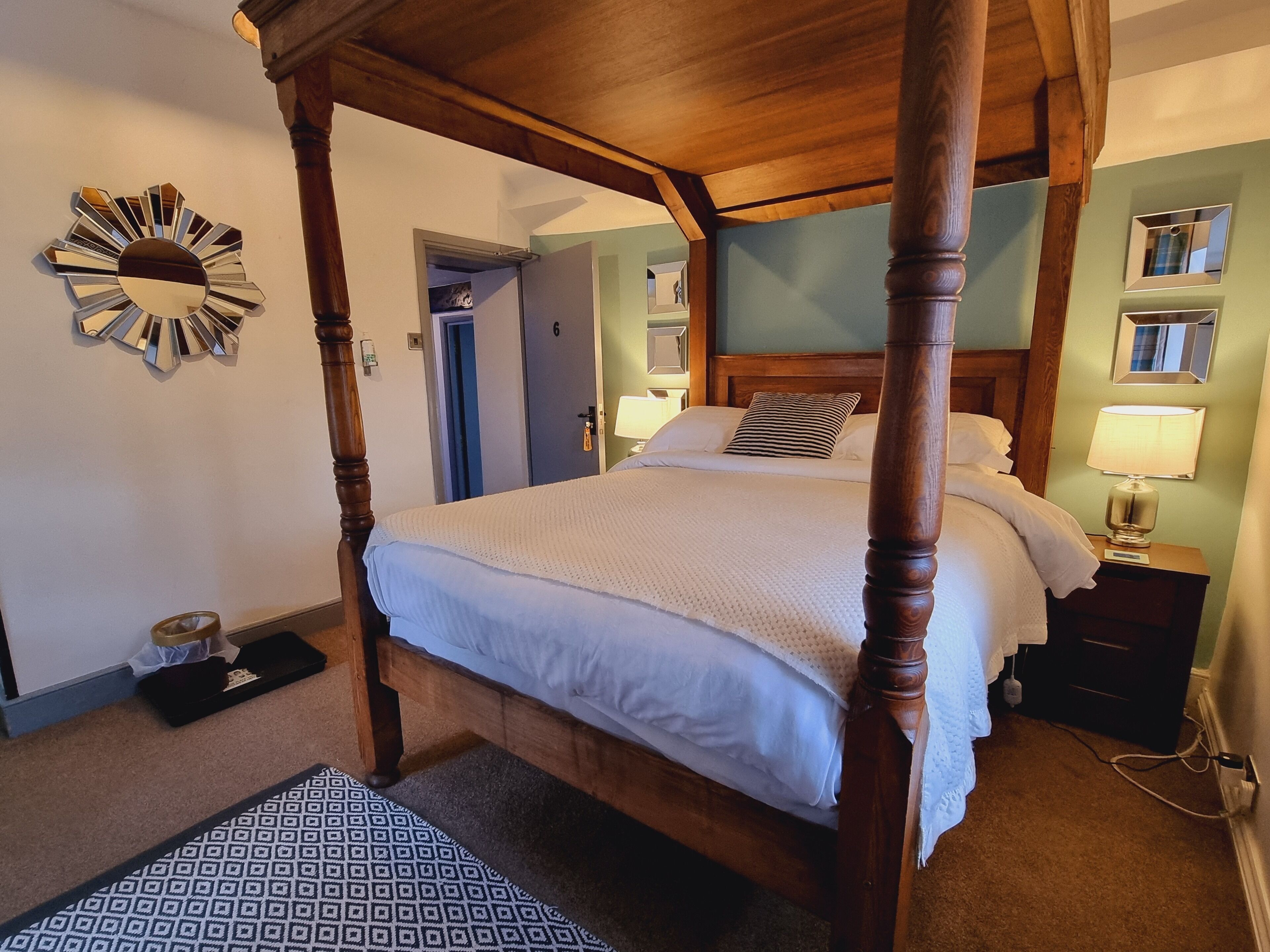 Een hemelbed met houten frame, een witte spreidek en een kamerbreed tapijt. Er staat een nachtkastje met een lamp, een spiegel aan de muur en een deur die leidt naar een andere kamer.