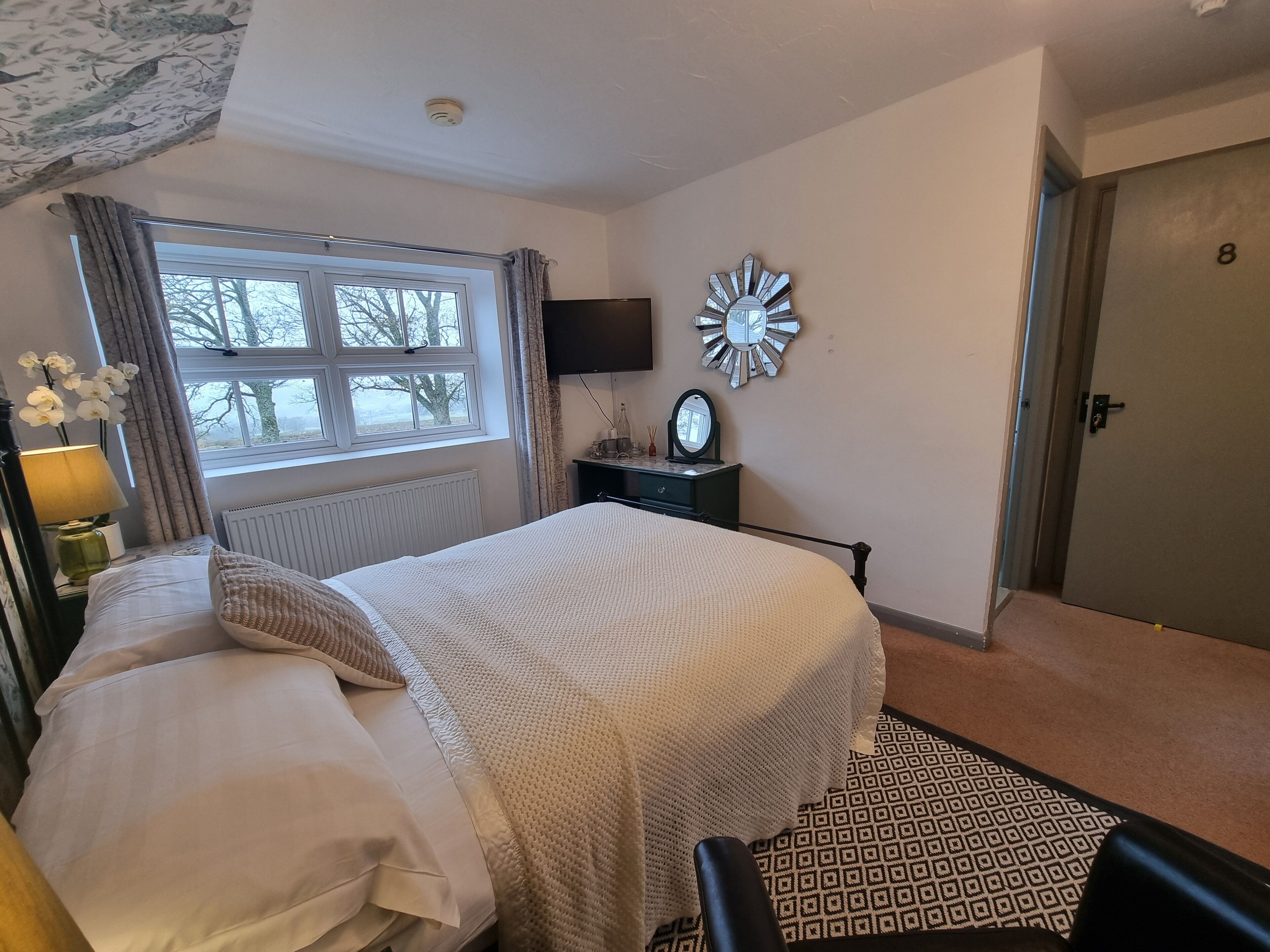 Un dormitorio con cama, mesita de noche, espejo, reloj de pared y puerta.