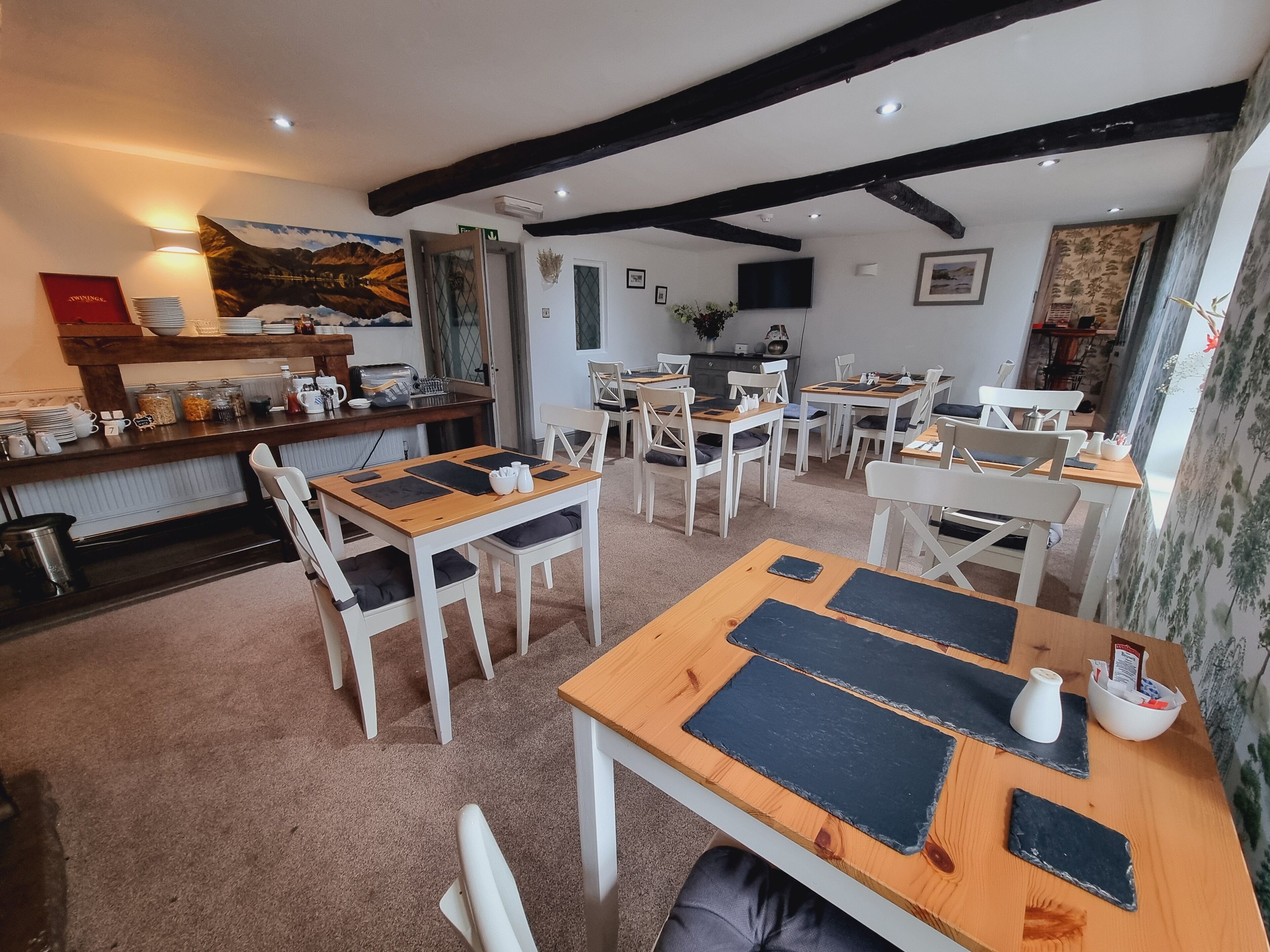 A cozy dining area with wooden tables and white chairs, a kitchen in the background, and a mountain painting on the wall.