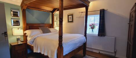 A four-poster bed with a wooden frame, a bedside table with a lamp, a mirror, and a framed picture on the wall.