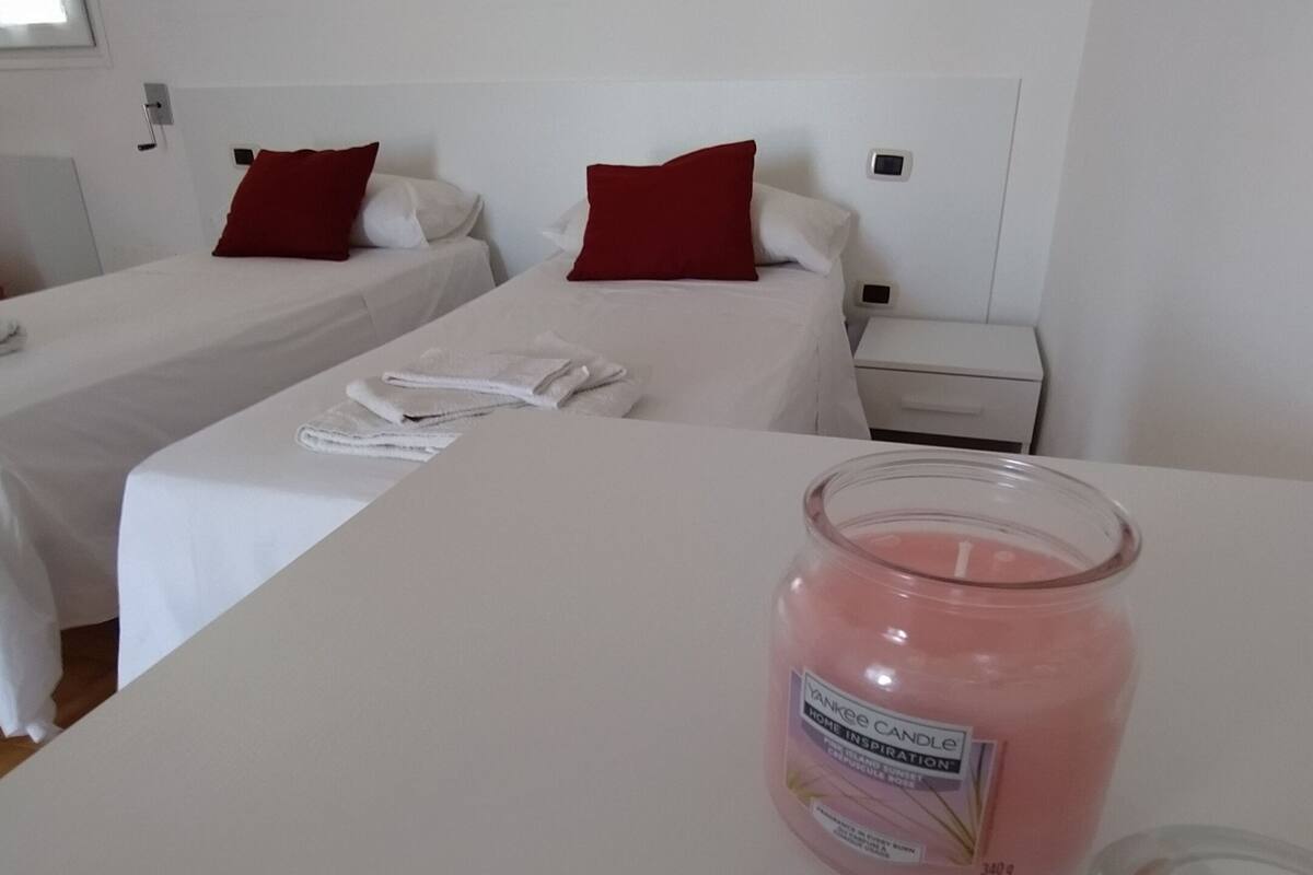 Habitación de hotel con dos camas, ropa blanca, almohadas rojas, una mesita de noche con una vela rosa y una mesita auxiliar.