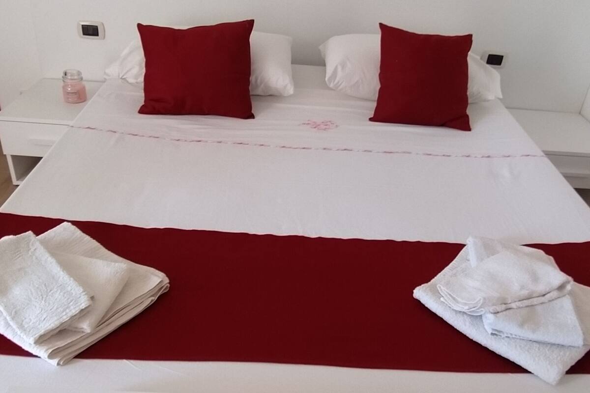 Una cama con ropa blanca y almohadas rojas, una mesita de noche blanca con una botella rosa, y dos toallas blancas dobladas sobre la cama.