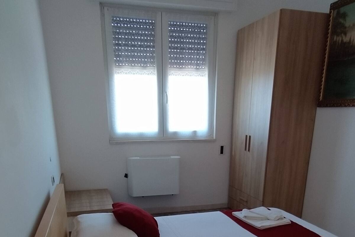 Un dormitorio con una cama grande, dos almohadas rojas, un edredón blanco, un armario de madera y una ventana con persianas.