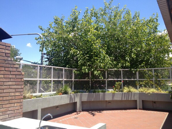 An outdoor patio area with a brick floor, a metal fence, and a large tree providing shade.