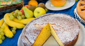 A round cake with a slice cut out, dusted with powdered sugar, surrounded by fruits like bananas, grapes, and apples. - Hotel Ervill (Rimini)