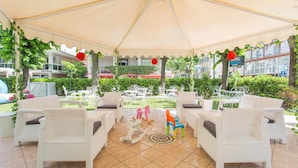 A covered outdoor seating area with white wicker furniture, a children's play area, and a view of the hotel and surrounding greenery. - Hotel Ervill (Rimini)