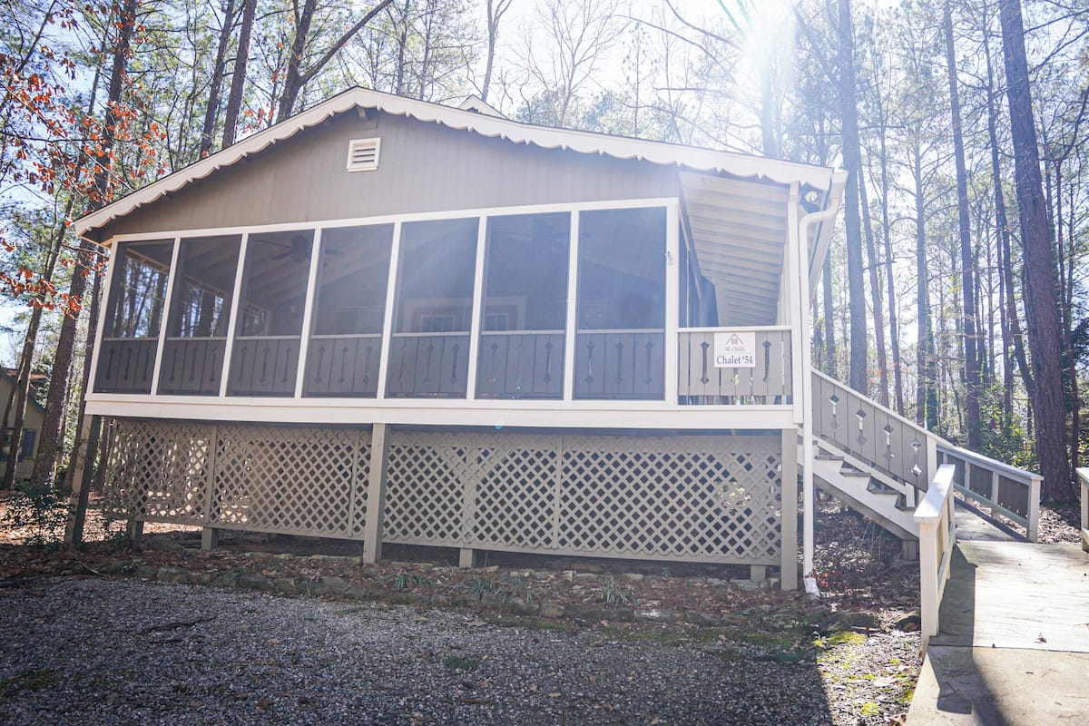 A cabin with a large windowed front, a wooden deck, and a staircase leading to the entrance.