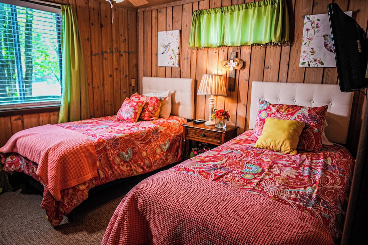 A room with two beds, a nightstand, a lamp, and a window with curtains.