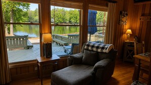 A cozy cabin with a wooden interior, a view of a lake, a chair, and a table with a lamp. - Pine Mountain Club Chalets Resort (Pine Mountain)