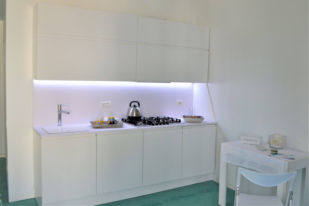 Cocina compacta con muebles blancos, estufa a gas, tetera y una pequeña mesa con una silla.
