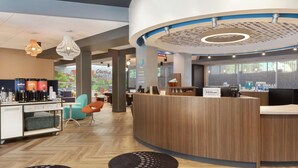Uma área de recepção de hotel moderna, com um balcão de madeira, uma estação de café e um espaço de estar com cadeiras.