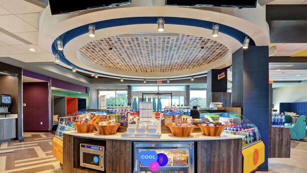 A modern hotel lobby with a central counter, a refrigerated display, and a seating area. - Tru by Hilton Baton Rouge Citiplace, LA (Baton Rouge)