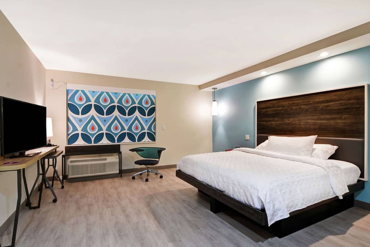 A hotel room with a bed, a desk with a TV, a chair, and a wall-mounted air conditioner.