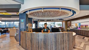 A modern hotel reception area with a curved wooden counter, a large window offering a view of the outside, and a visible Hilton logo. - Tru by Hilton Baton Rouge Citiplace, LA (Baton Rouge)