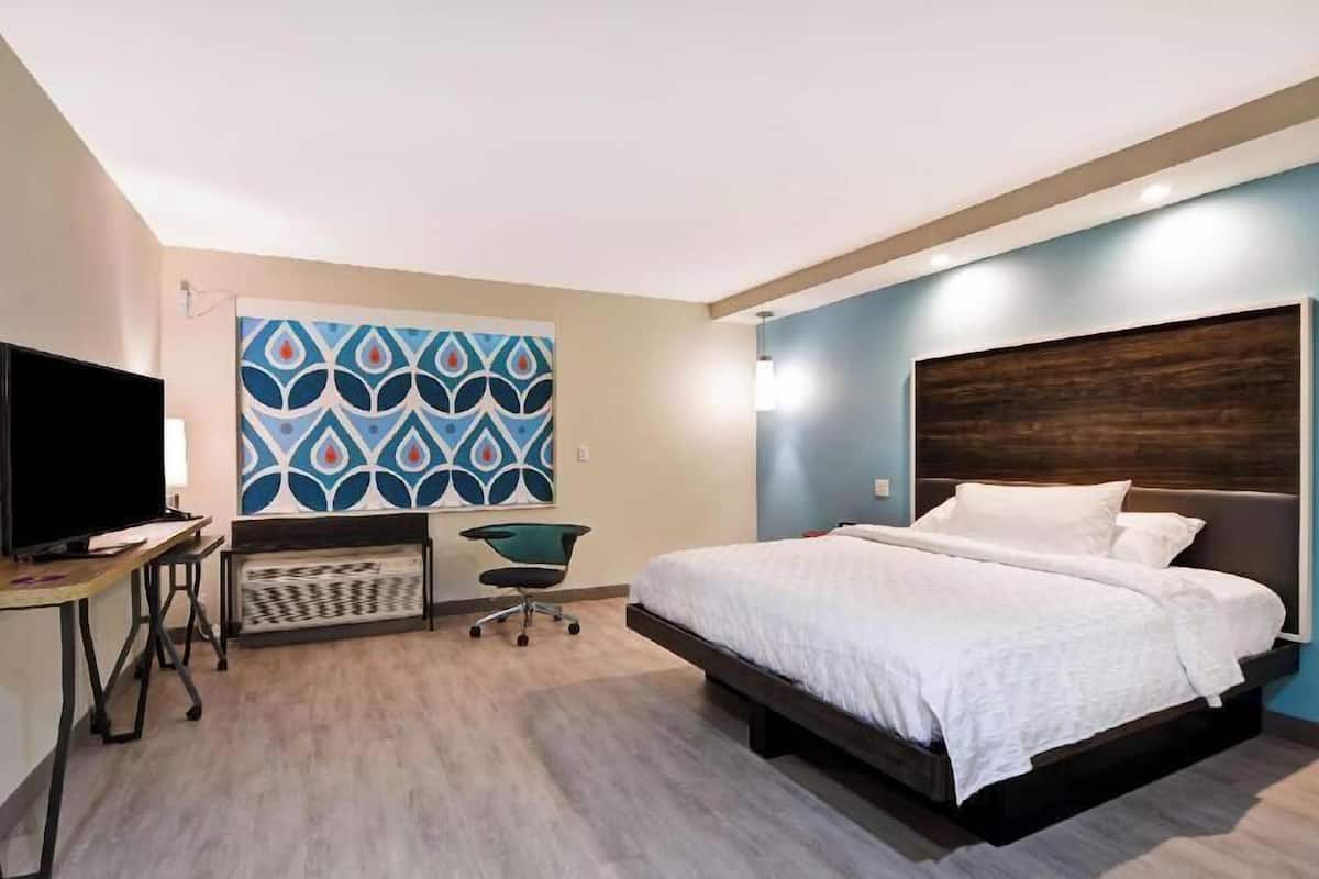 A modern hotel room with a bed, a desk with a flat-screen TV, a chair, and a decorative wall pattern.
