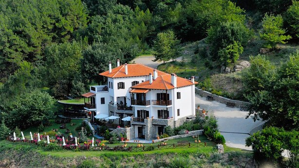 A multi-story white building with a red-tiled roof, surrounded by lush greenery and a paved pathway leading to a pool. - Nastou View Hotel (Sintiki)