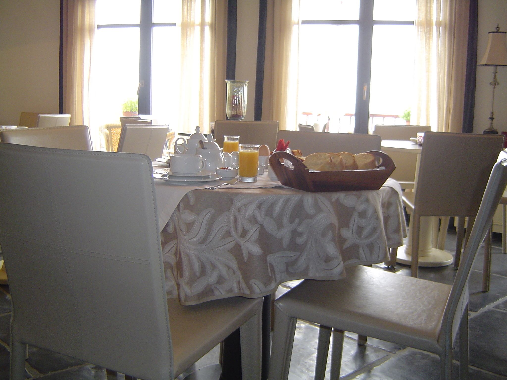 A dining area with a table set for breakfast, including a basket of bread, a teapot, cups, and a glass of orange juice.