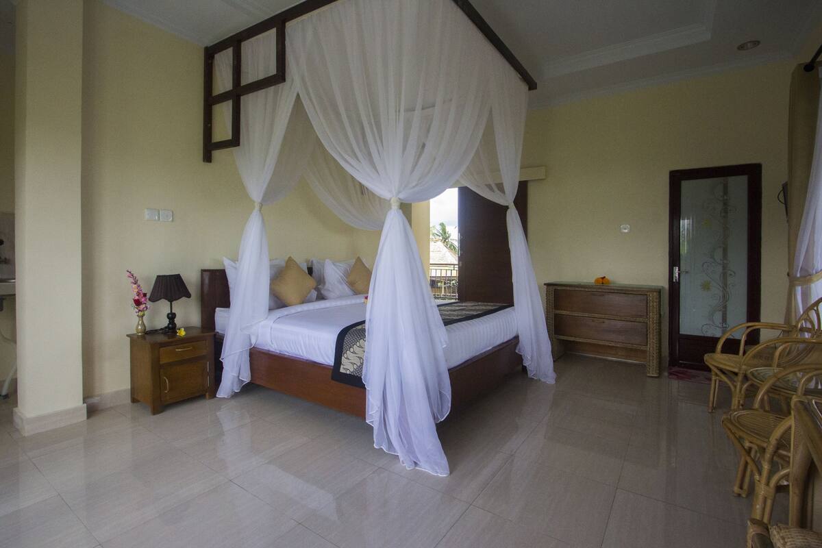 A bedroom with a canopy bed, a nightstand with a lamp, a dresser, and wicker chairs.