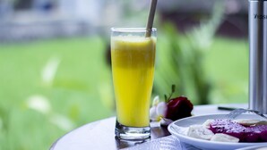 A glass of yellow juice with a straw, a plate with food, and a thermos on a table with a green outdoor background. - Purnama Guest House (Ubud)