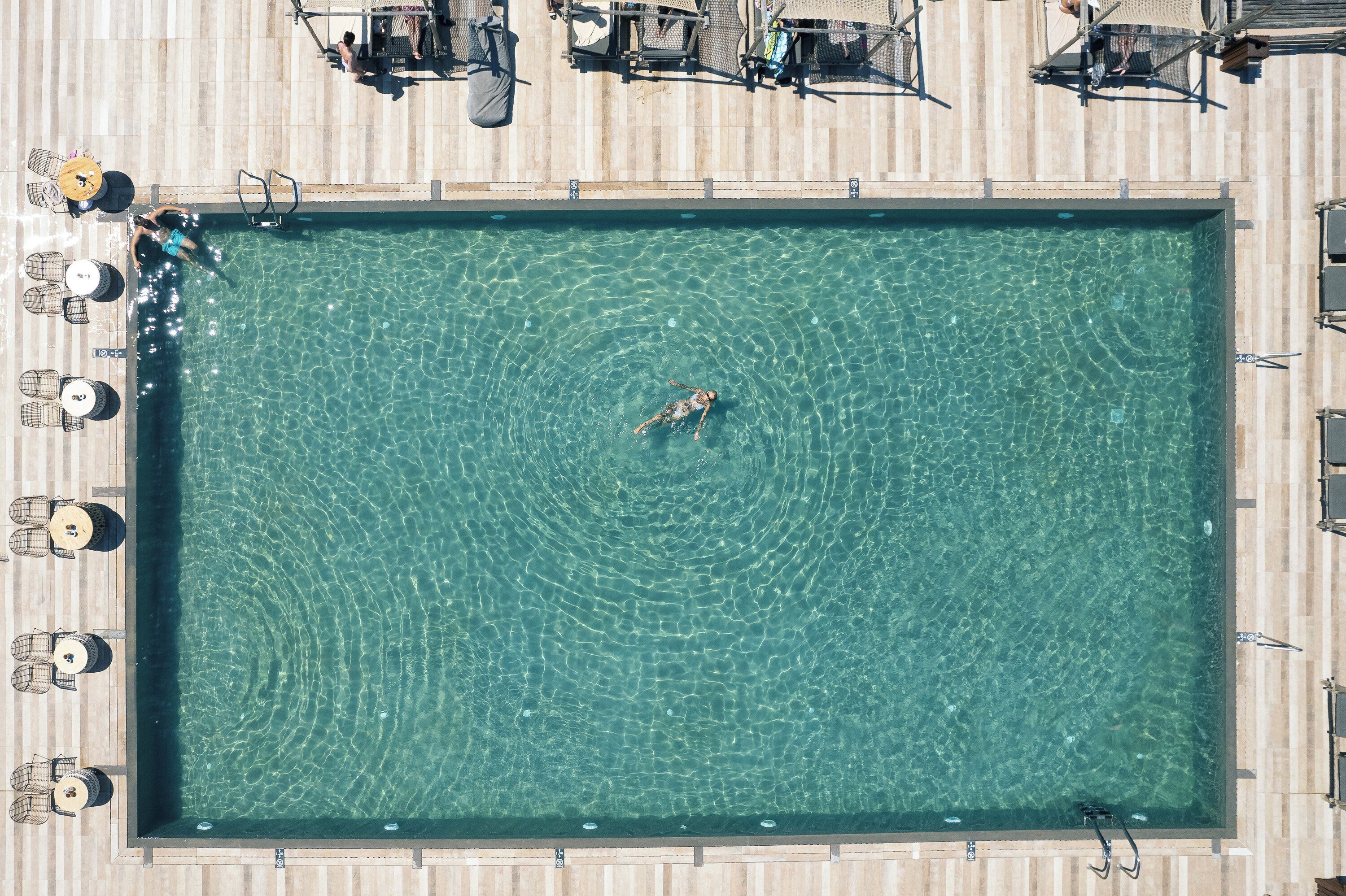 Une vue aérienne d’une piscine rectangulaire à l’eau claire, entourée de chaises longues et de parasols.