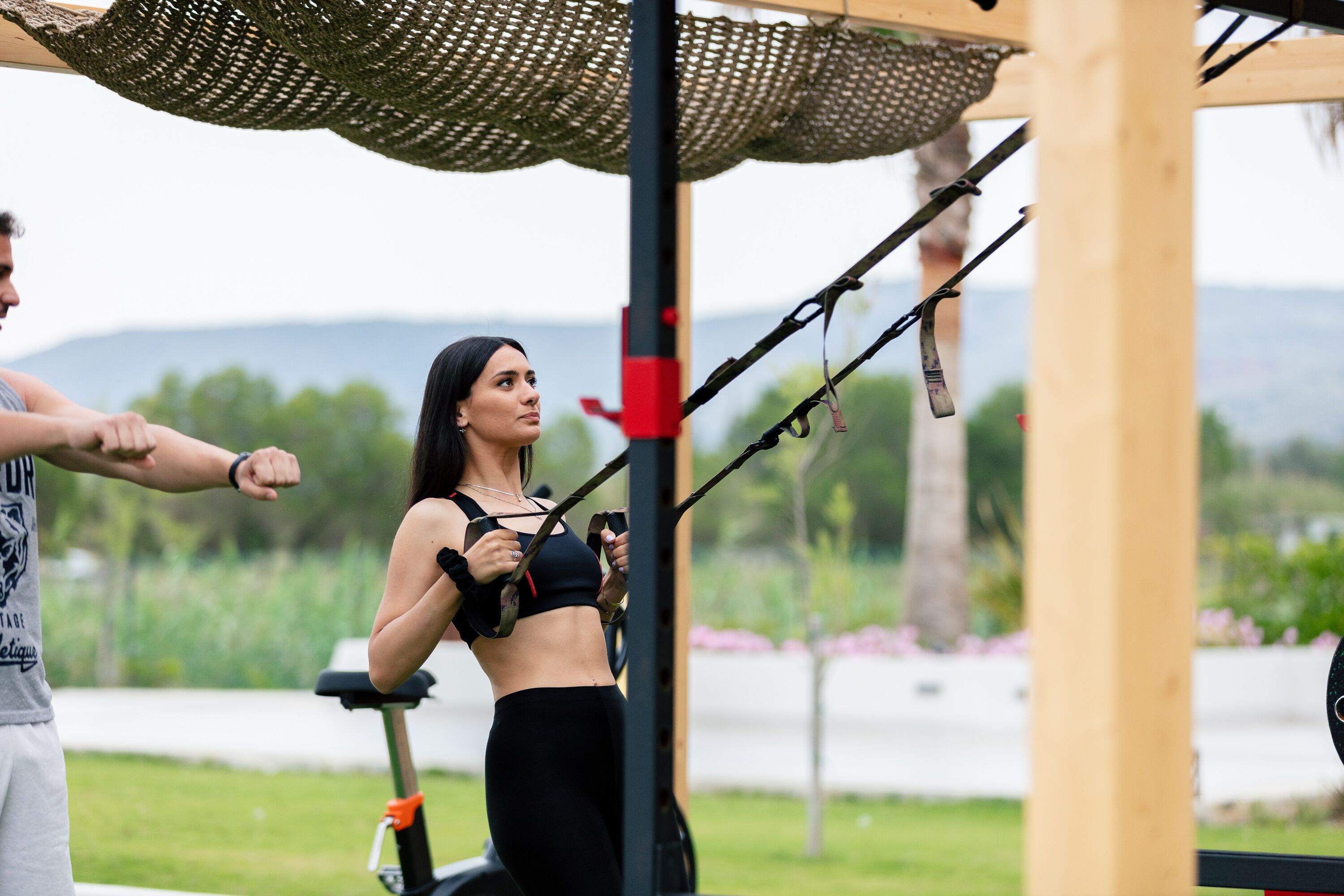 A person using a suspension trainer in an outdoor gym area with a view of a lake and hills in the background.