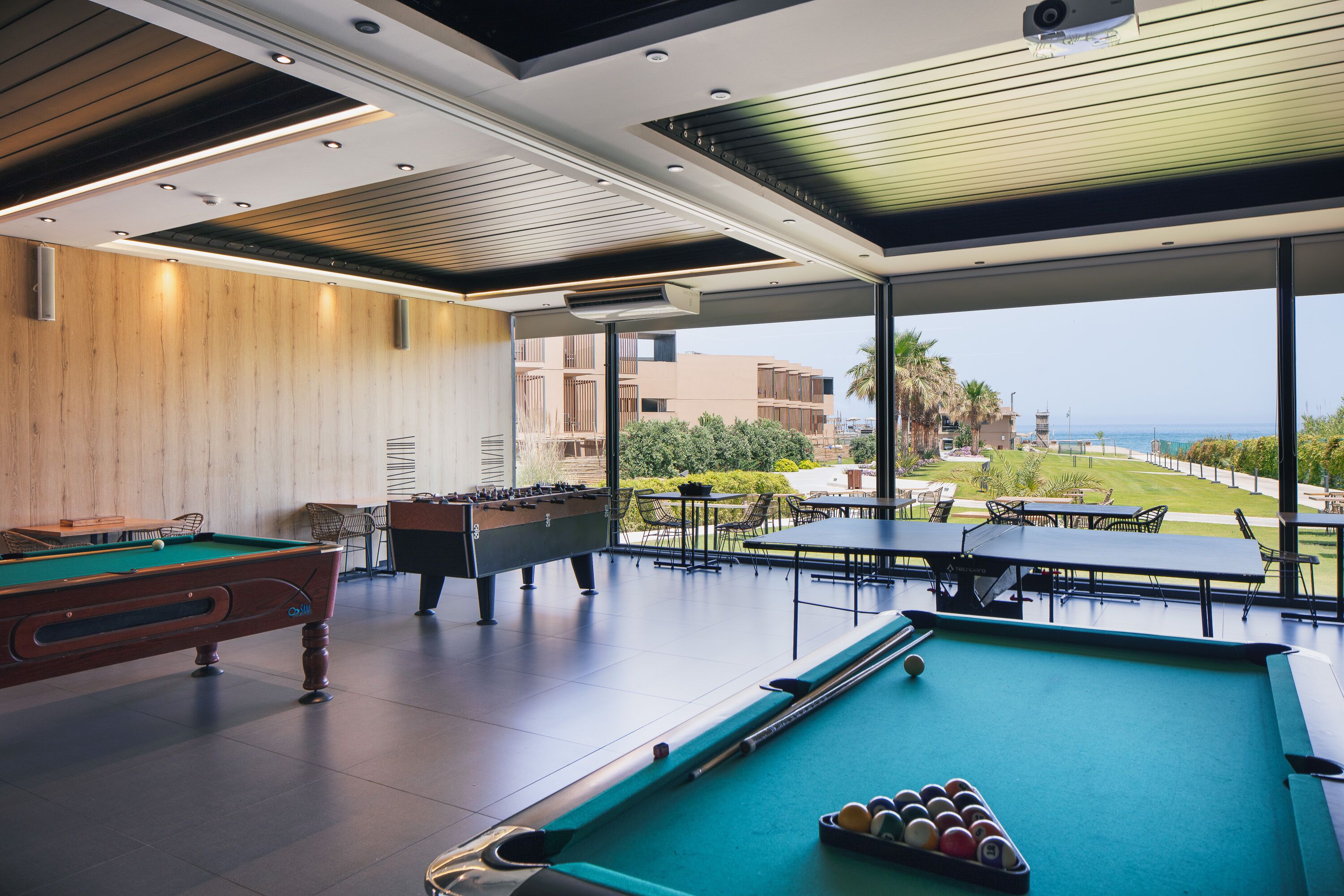 Zona de piscina con mesa de fútbol, mesa de billar y bolas de billar, con vistas a un jardín ajardinado y edificios.