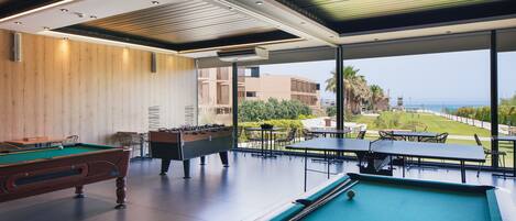 A pool area with a foosball table, pool table, and billiard balls, overlooking a landscaped garden and buildings.
