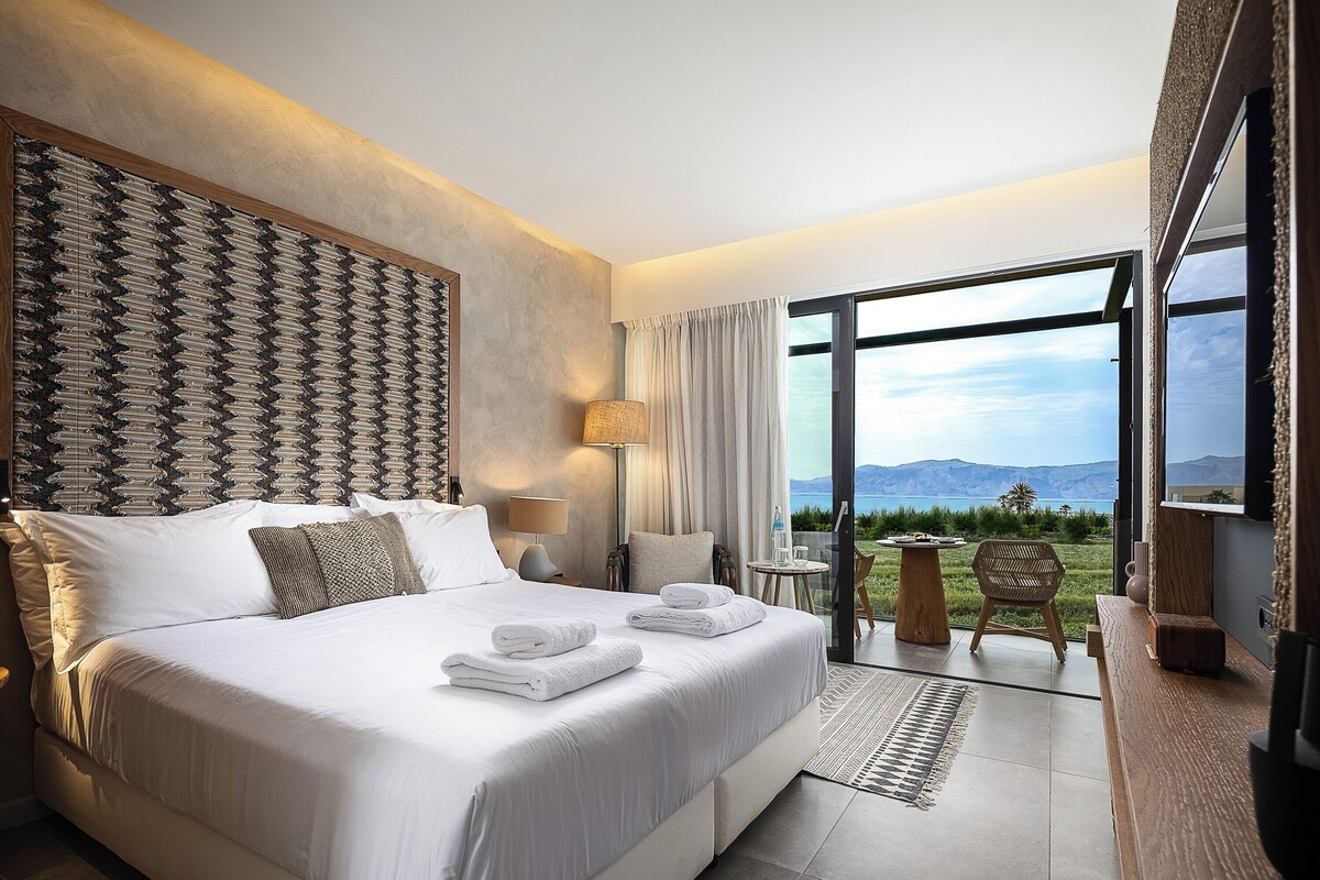 A modern hotel room with a large bed, a view of the outdoors, and a small seating area.