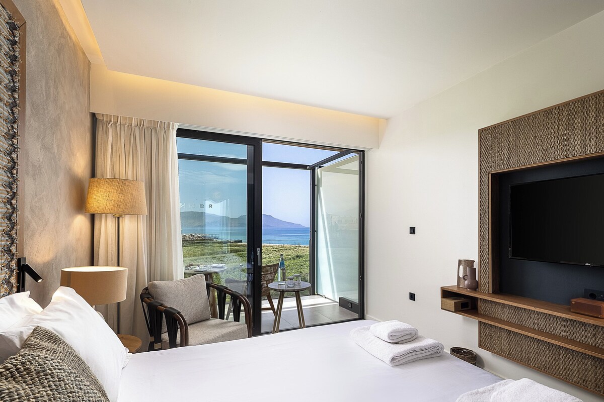 A hotel room with a bed, a chair, a table, a TV, and a view of the sea.