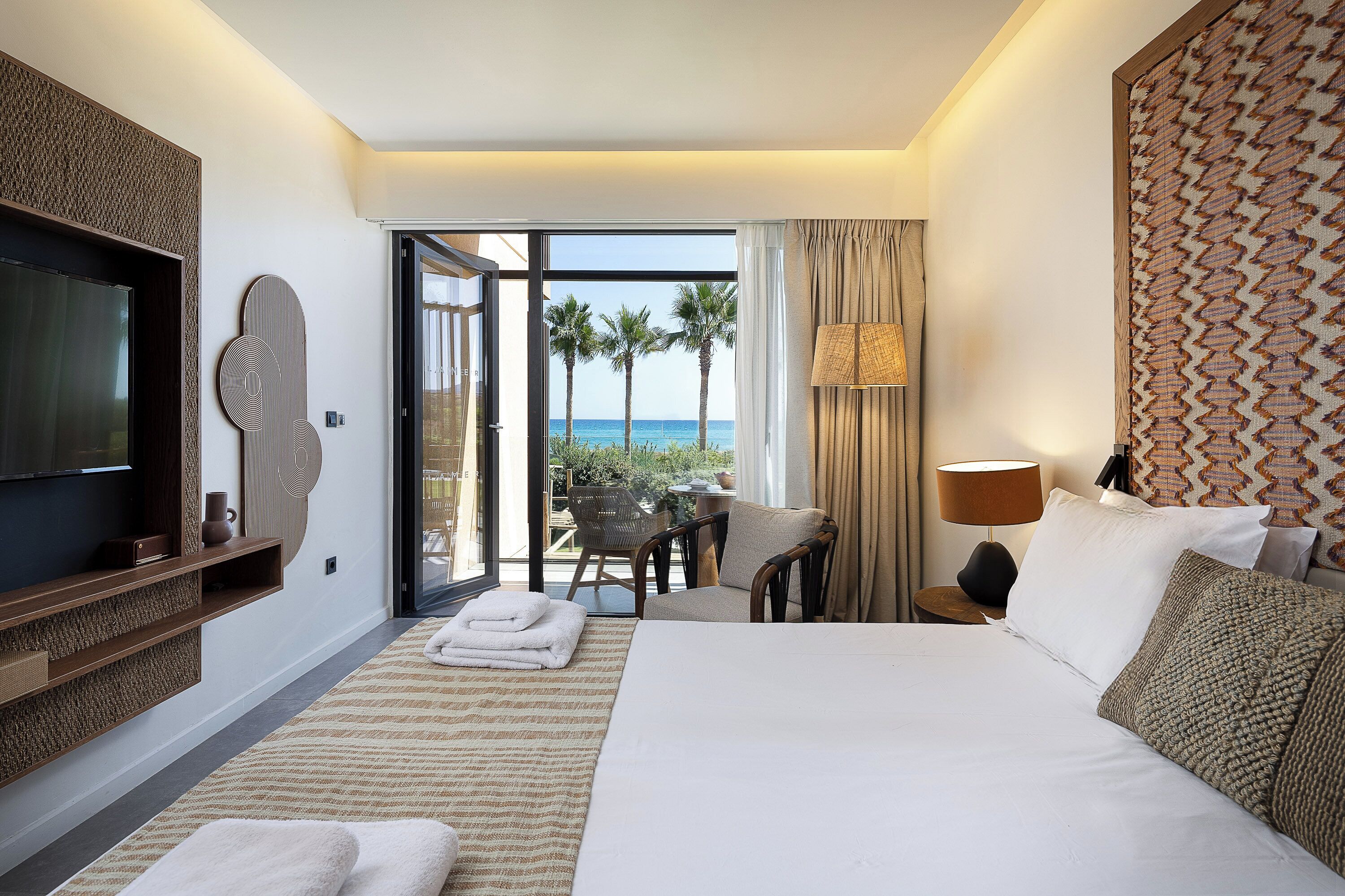 Camera d'albergo con un letto grande, televisione, un balcone vista palme e mare, e un'area ristorazione con tavolo e sedie.