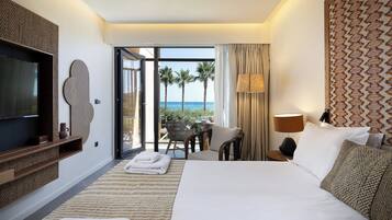 Camera d'albergo con un letto grande, televisione, un balcone vista palme e mare, e un'area ristorazione con tavolo e sedie.