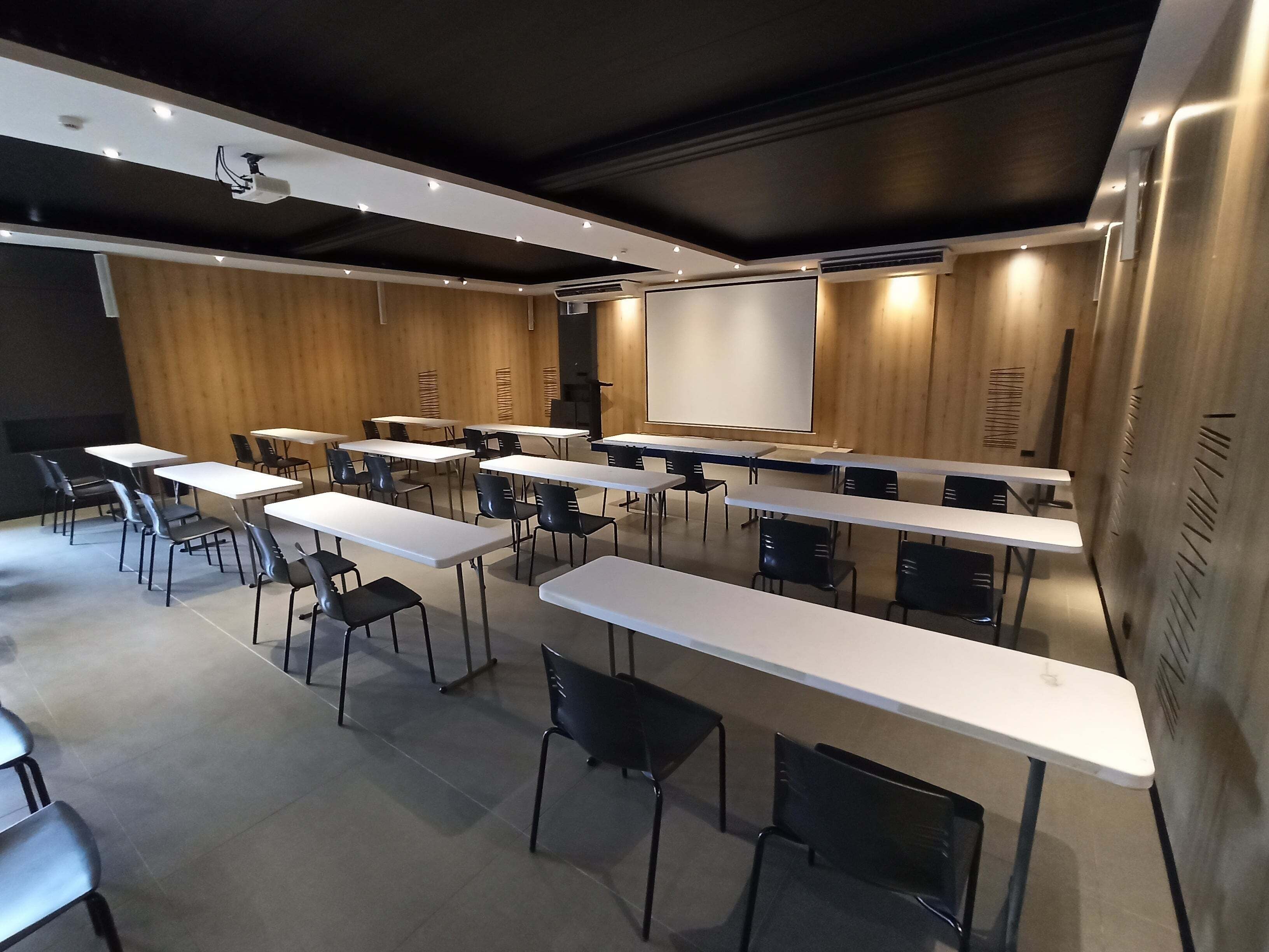 A conference room with white tables and black chairs, a large screen, and wooden panel walls.