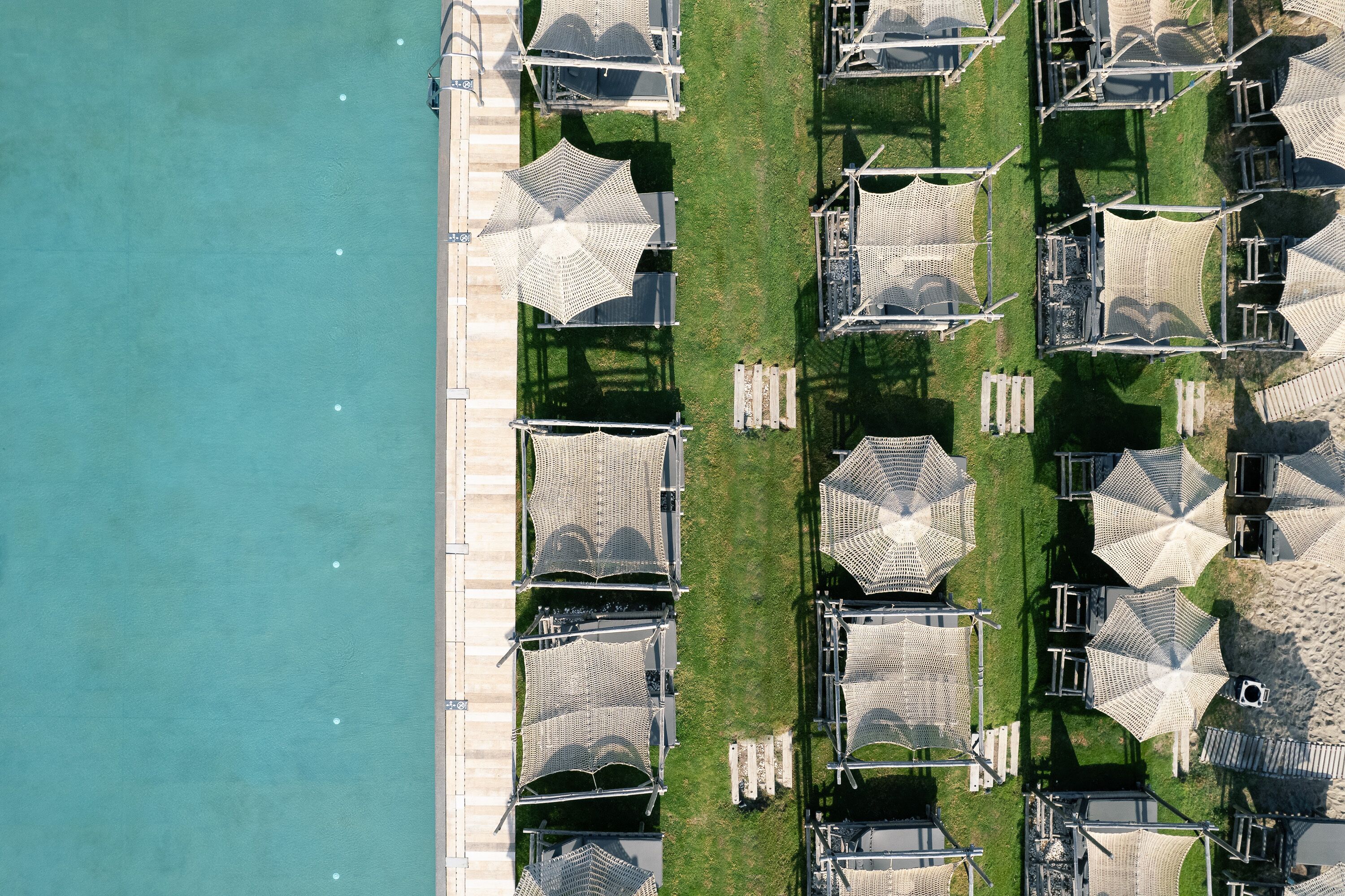 Vue aérienne d’un complexe hôtelier en bord de mer, avec des rangées de parasols et de chaises longues alignées parallèlement au rivage.
