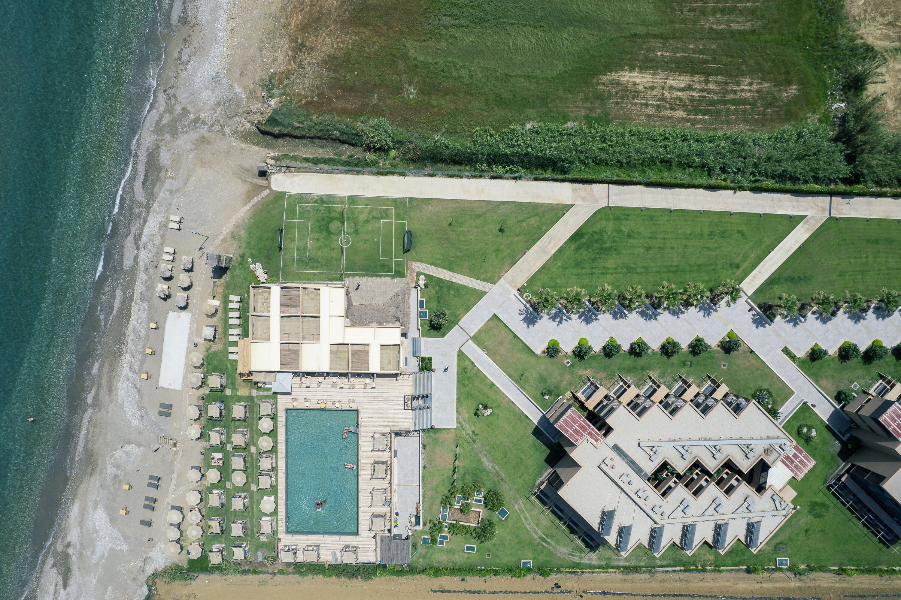 An aerial view of a coastal hotel with a swimming pool, beachside seating, and a tennis court.