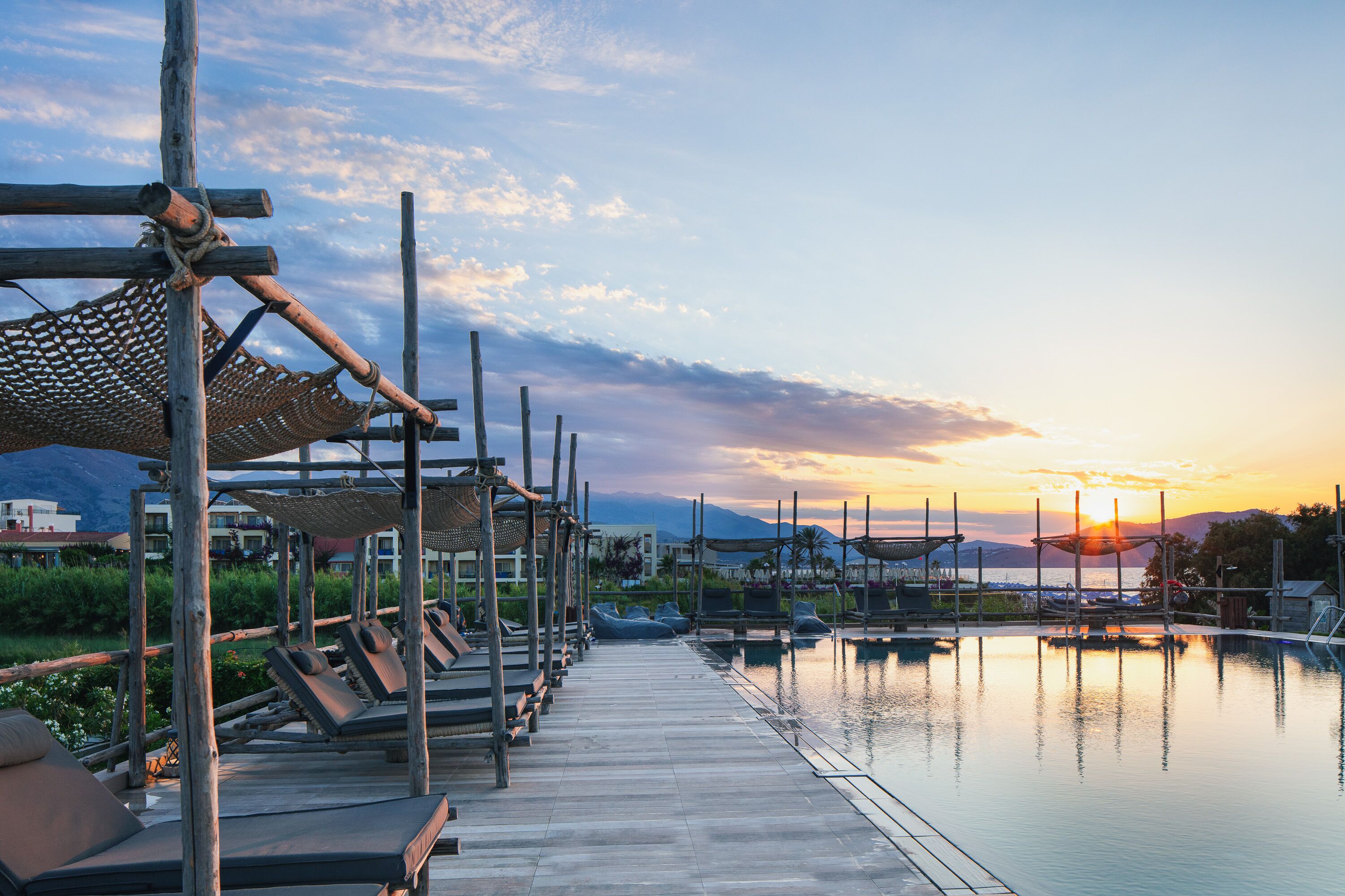 Une terrasse en bois avec des chaises longues et une piscine, le tout baigné par les couleurs du coucher de soleil.