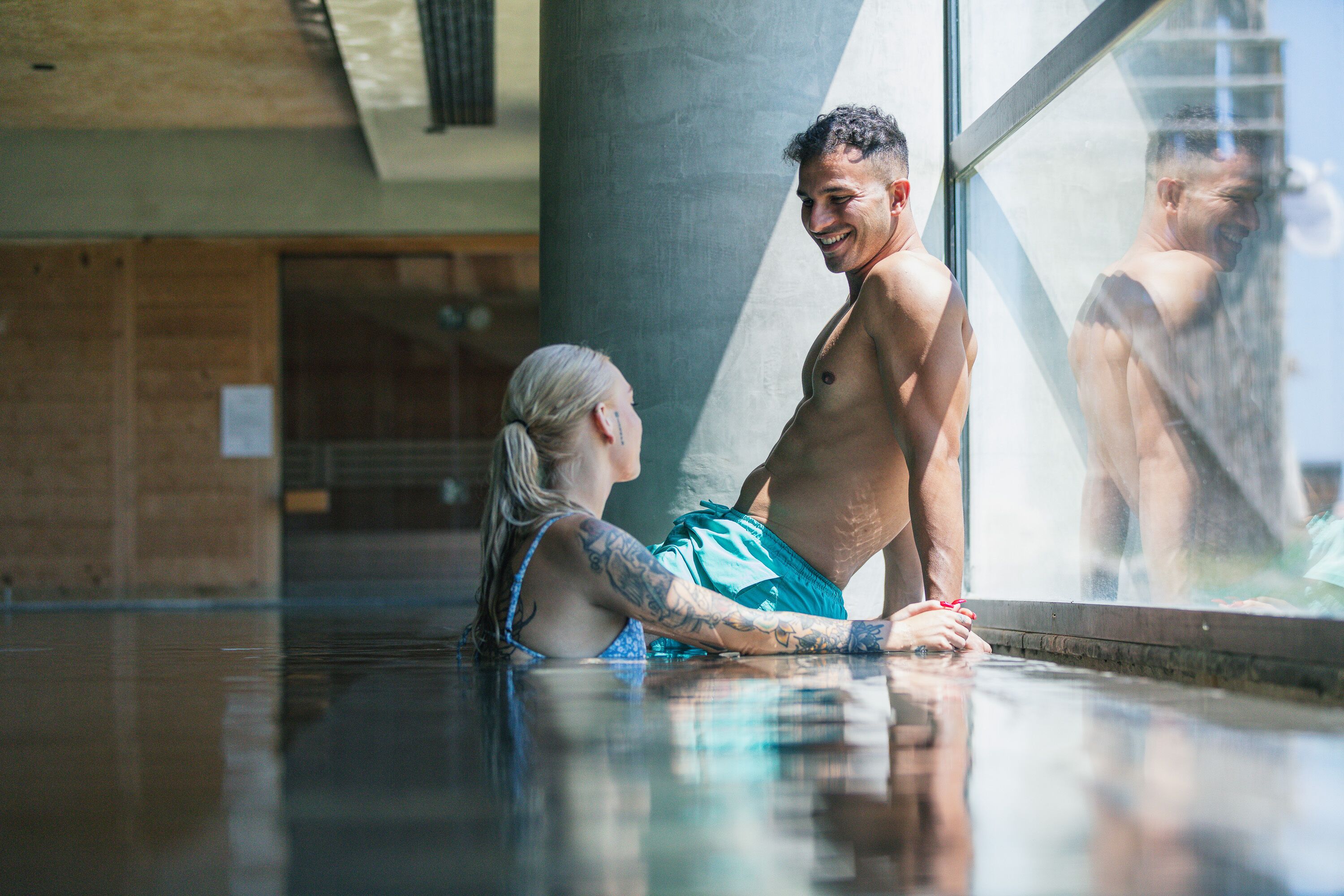 Un hombre y una mujer en una piscina con un amplio ventanal que refleja su imagen.