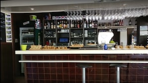 A bar area with shelves of bottles and glasses, a computer monitor, and a counter with food items. - Hostal Isla Playa (Arnuero)