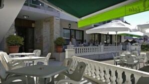 An outdoor café with white plastic tables and chairs, a green awning, and a white railing. - Hostal Isla Playa (Arnuero)