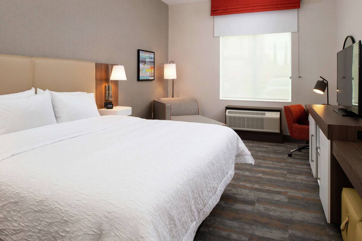 Une chambre d’hôtel avec un grand lit, un bureau avec un ordinateur, une chaise, une télévision et une fenêtre agrémentée d’un rideau rouge.